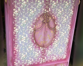 Può includere: Un armadio in stile vintage con una finitura rosa rovinata e dettagli dorati. Le porte presentano un motivo floreale e un design ovale decorativo. L'armadio ha una sezione inferiore con tre cassetti.