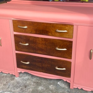 May include: A vintage-style coral-colored buffet with three central drawers and two side cabinets. The drawers are a dark wood, and the handles are gold. The buffet has a scalloped base and gold trim.