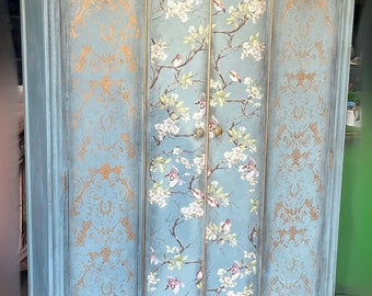 May include: A three-door armoire with a distressed blue paint finish and gold accents. The doors feature a floral design with birds and a lace pattern.