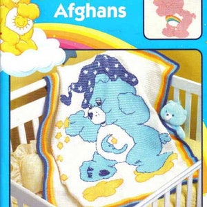 May include: A crochet pattern book titled "Care Bears Afghans" features a blue Care Bear design on a white blanket with yellow and blue accents. The cover is bright blue with a rainbow and a yellow Care Bear illustration.
