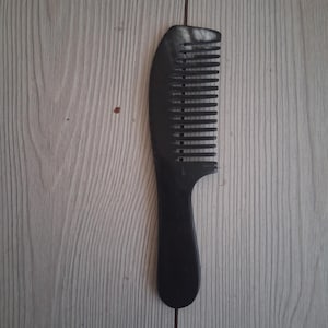 Könnte beinhalten: Ein schwarzer Haarkamm aus Kunststoff mit einem soliden, abgerundeten Griff und einer Reihe breiter, gleichmäßig angeordneter Zinken. Der Kamm hat eine glatte, matte Oberfläche und ein geschwungenes Design, das sich vom Griff zur Spitze verjüngt.
