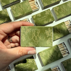 May include: A rectangular matchbox with a green cover featuring a gold floral pattern and a gold monogram. The text reads Claire & Michael, New Orleans, Louisiana, April 18, 2024. The matchbox contains wooden matches with white tips.
