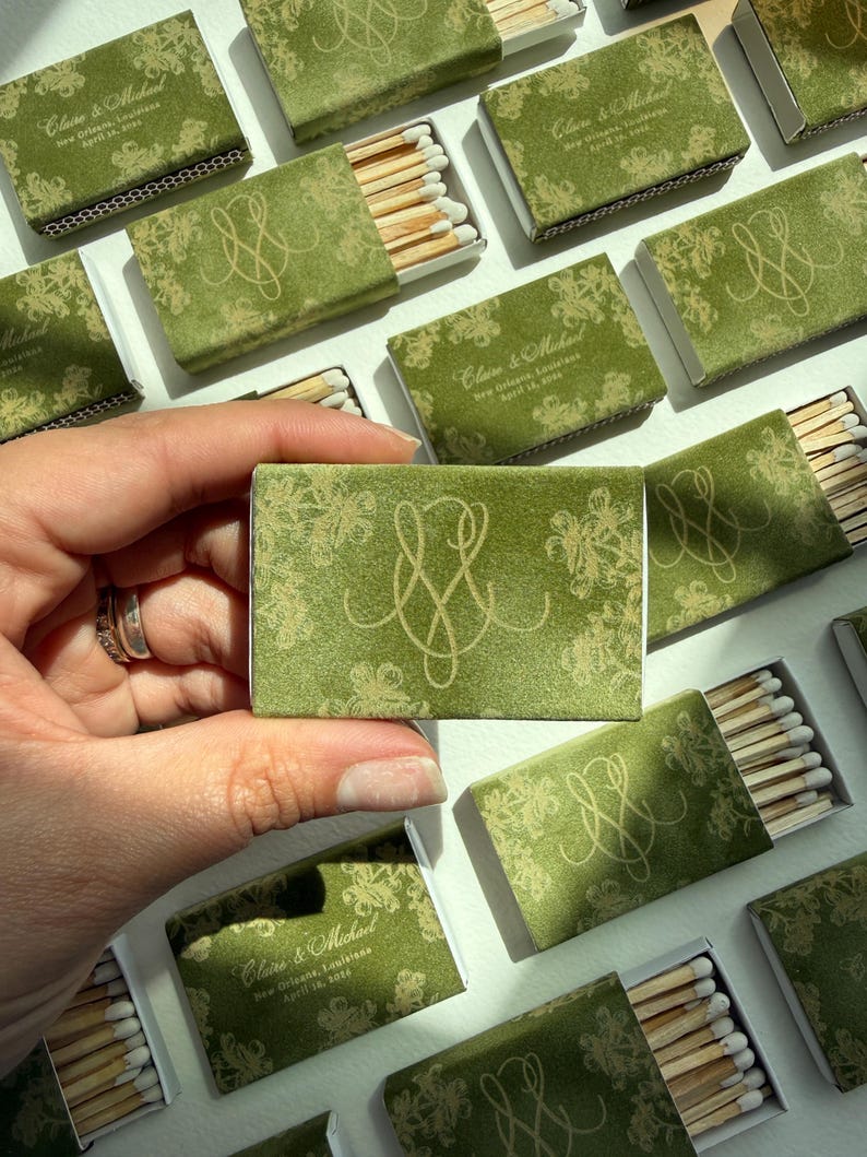 May include: A rectangular matchbox with a green cover featuring a gold floral pattern and a gold monogram. The text reads Claire & Michael, New Orleans, Louisiana, April 18, 2024. The matchbox contains wooden matches with white tips.