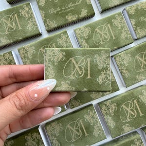 May include: A collection of rectangular matchboxes covered in olive green paper with a gold floral pattern. Each box features a gold monogram of the letters M and M, while some display the text Baylee & Marshall perfect match in gold.