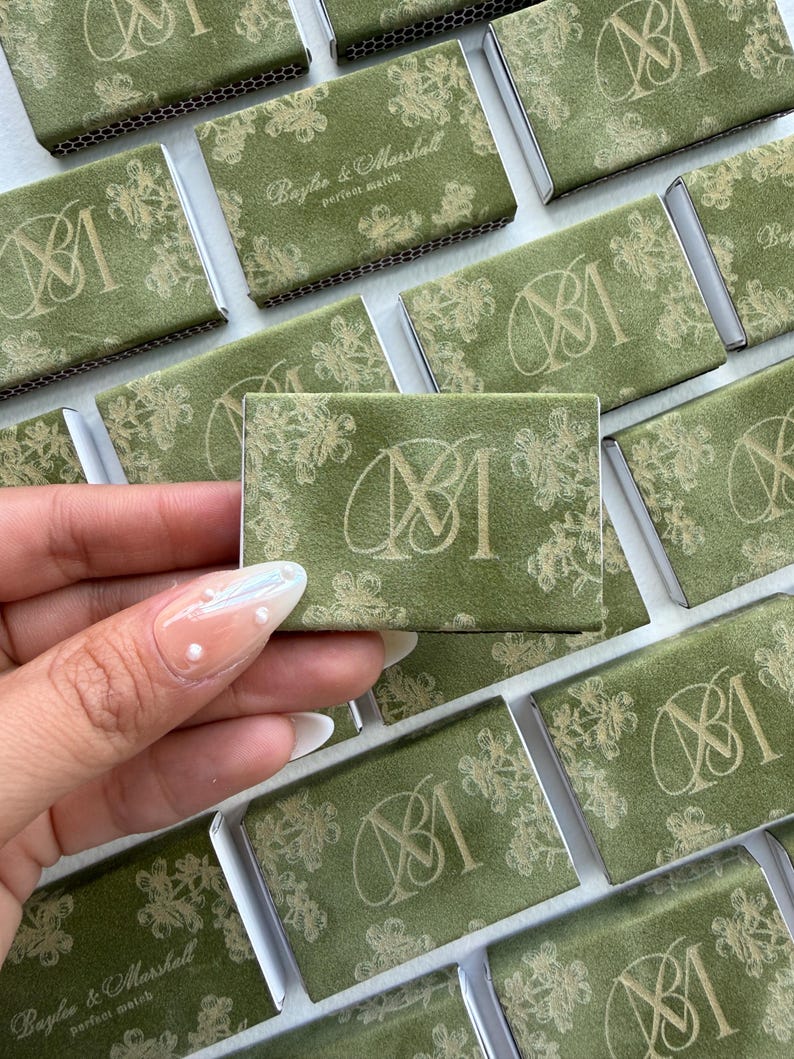 May include: A collection of rectangular matchboxes covered in olive green paper with a gold floral pattern. Each box features a gold monogram of the letters M and M, while some display the text Baylee & Marshall perfect match in gold.