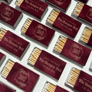 May include: A collection of rectangular matchboxes with a deep burgundy velvet-like finish. Each box features gold foil text reading "SAVE THE DATE The Vampire Ball 10.30.26" or a decorative gold monogram letter M surrounded by floral filigree.