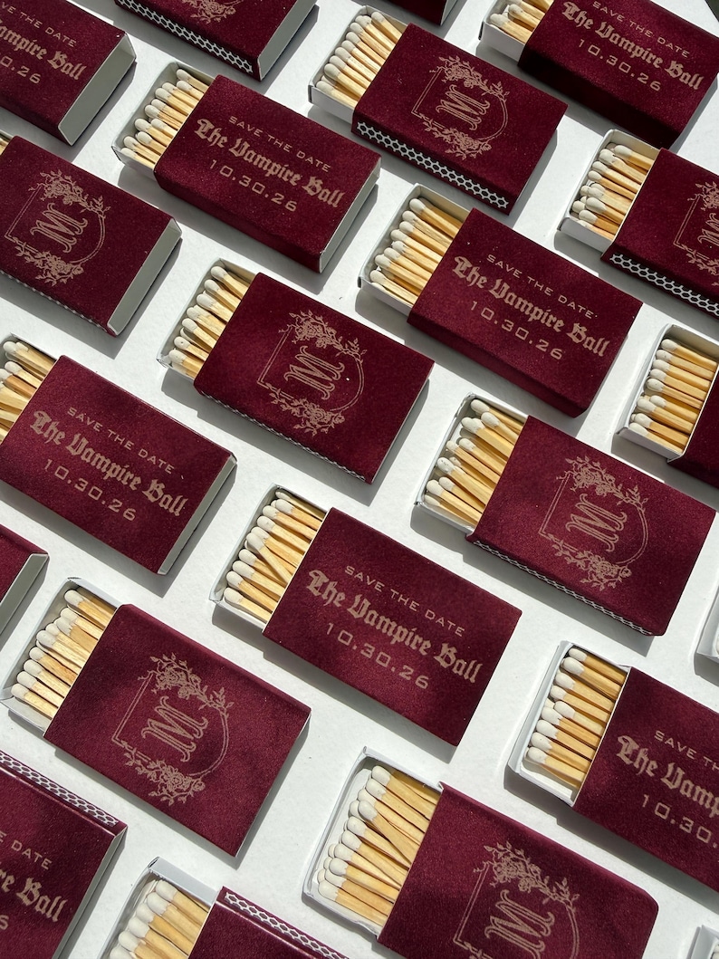 May include: A collection of rectangular matchboxes with a deep burgundy velvet-like finish. Each box features gold foil text reading "SAVE THE DATE The Vampire Ball 10.30.26" or a decorative gold monogram letter M surrounded by floral filigree.