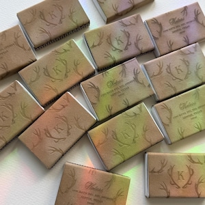 May include: A collection of rectangular matchboxes featuring a gradient of tan, green, and purple hues. Each box displays an embossed letter K centred within a pair of antlers, with the text Katari 1775 Gato Del Sol Avenue Austin, Texas 78738.