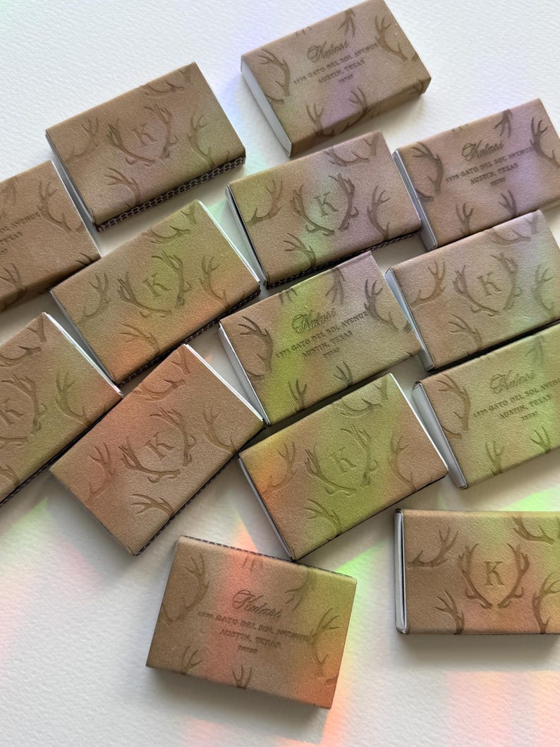 May include: A collection of rectangular matchboxes featuring a gradient of tan, green, and purple hues. Each box displays an embossed letter K centred within a pair of antlers, with the text Katari 1775 Gato Del Sol Avenue Austin, Texas 78738.