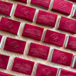 May include: A collection of rectangular matchboxes covered in deep red velvet. Some feature a gold-toned line drawing of Dundas Castle, Edinburgh, Scotland, while others display the gold-toned cursive initials CR and the date September 4, 2026.