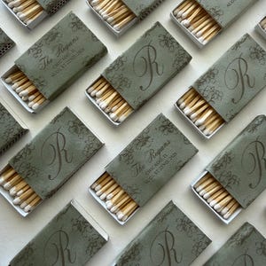 May include: Multiple rectangular matchboxes with sage green covers featuring a dark green floral pattern and a large letter R. Some boxes display the text The Regans, Chicago, IL, August 22nd, 2026. Each box is partially open, revealing wooden matches.