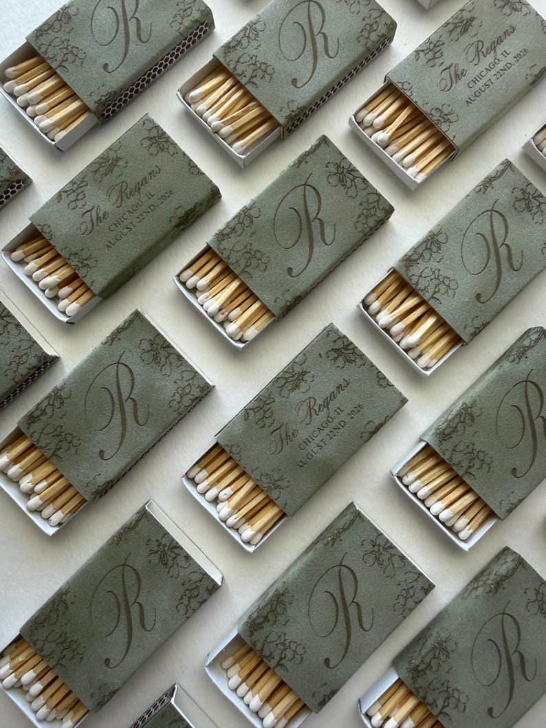 May include: Multiple rectangular matchboxes with sage green covers featuring a dark green floral pattern and a large letter R. Some boxes display the text The Regans, Chicago, IL, August 22nd, 2026. Each box is partially open, revealing wooden matches.