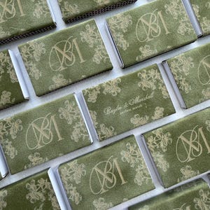 May include: A collection of rectangular matchboxes covered in olive green textured paper. Each box features a gold foil floral pattern and either a monogram of the letters BM or the text Baylee & Marshall perfect match printed in gold.