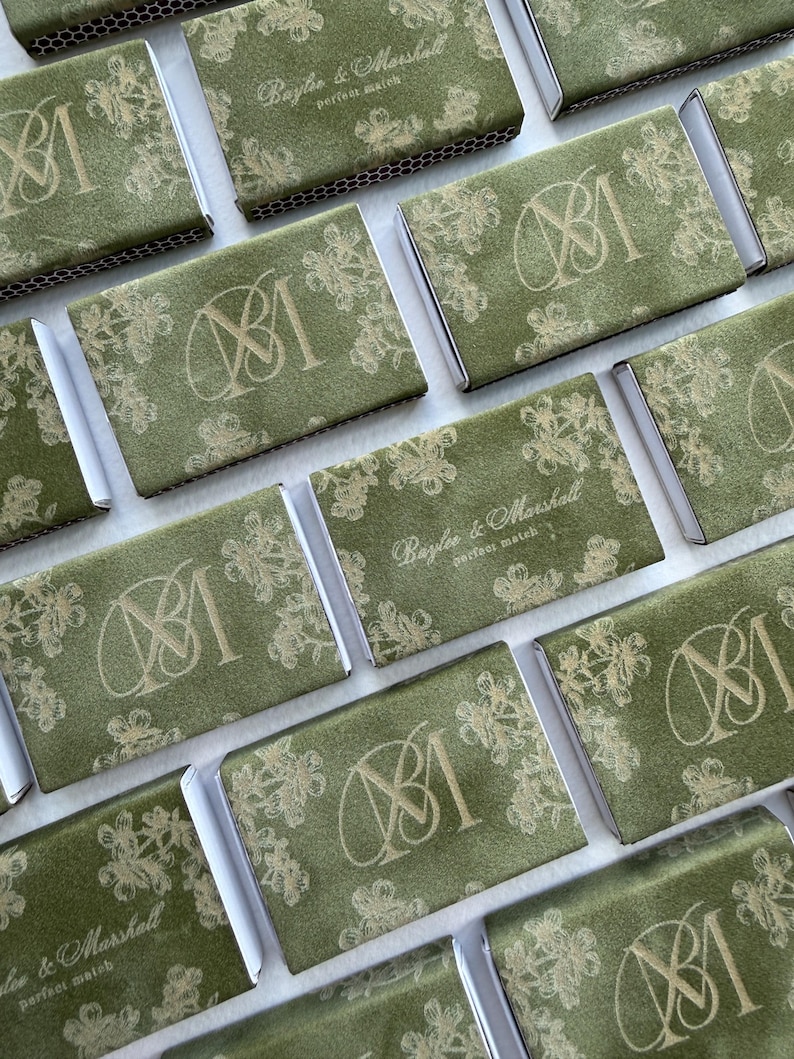 May include: A collection of rectangular matchboxes covered in olive green textured paper. Each box features a gold foil floral pattern and either a monogram of the letters BM or the text Baylee & Marshall perfect match printed in gold.