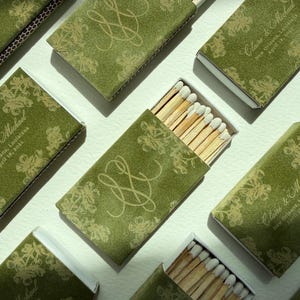 May include: Olive green matchboxes with floral designs and custom text. Some boxes are open, revealing wooden matches with white tips. The boxes feature a monogram and names with a date.