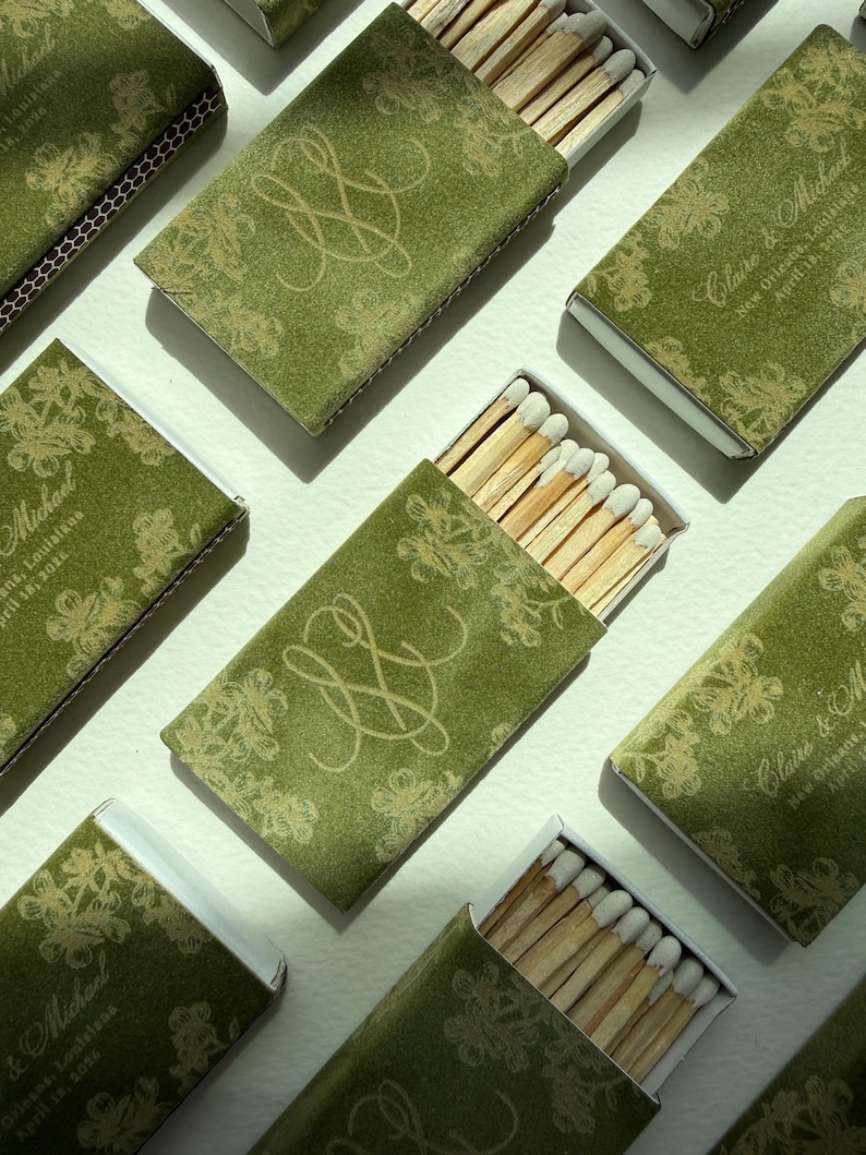 May include: Multiple rectangular matchboxes featuring a moss green cover with a gold floral pattern and a gold monogram. Some boxes display the text Claire & Michael, New Orleans, Louisiana, April 18, 2024. Each box contains wooden matches with white tips.