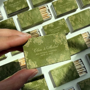 May include: A rectangular matchbox with a green cover featuring a gold floral pattern and the text Claire & Michael, New Orleans, Louisiana, April 18, 2026. The box contains wooden matches with white tips, arranged in a grid of multiple matchboxes.