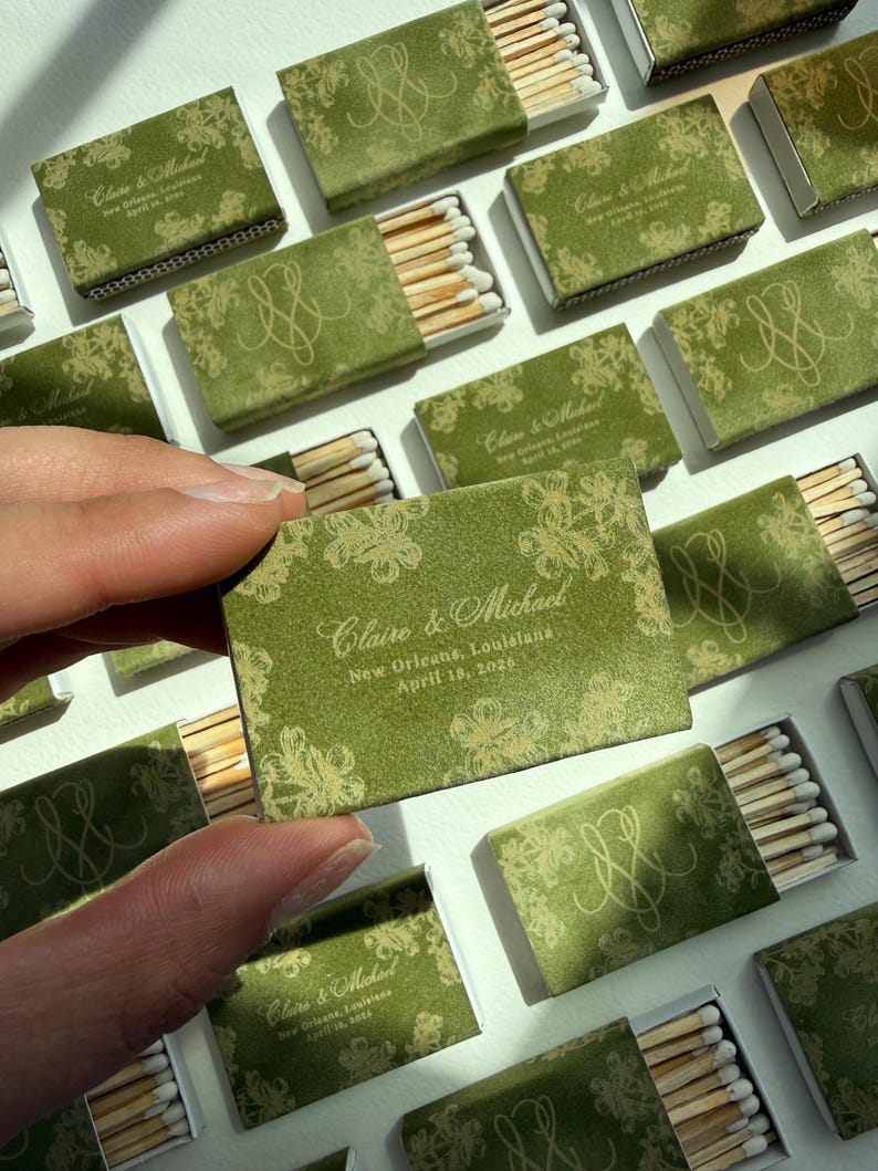 May include: A rectangular matchbox with a green cover featuring a gold floral pattern and the text Claire & Michael, New Orleans, Louisiana, April 18, 2026. The box contains wooden matches with white tips, arranged in a grid of multiple matchboxes.