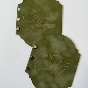 May include: Two olive green gift tags with decorative edges. Each tag has the word "TO" above a name in elegant script. One tag says "TO Julianne" and the other says "TO Nick". Each tag has three silver and burgundy rivets.