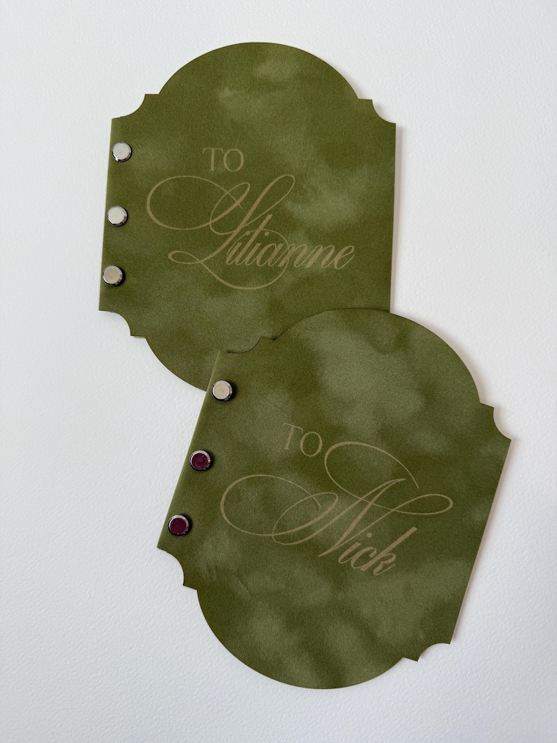 May include: Two olive green gift tags with decorative edges. Each tag has the word "TO" above a name in elegant script. One tag says "TO Julianne" and the other says "TO Nick". Each tag has three silver and burgundy rivets.