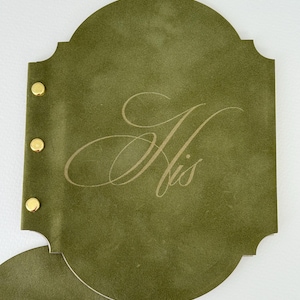 May include: Olive green leather-like journal with gold-colored rivets. The journal has a decorative shape and features the word "His" in an elegant, light-colored script. The journal is open, revealing a second page.