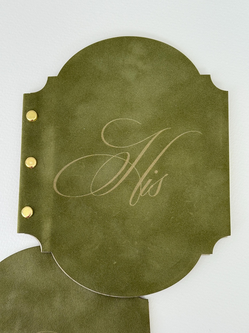 May include: Olive green leather-like journal with gold-colored rivets. The journal has a decorative shape and features the word "His" in an elegant, light-colored script. The journal is open, revealing a second page.