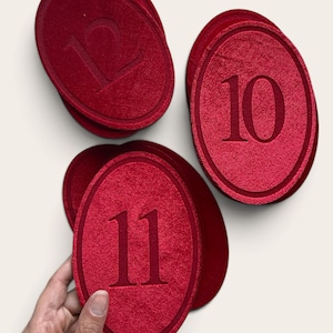K&ouml;nnte beinhalten: Ein Set aus ovalen, roten Stoffanh&auml;ngern mit den gepr&auml;gten Zahlen 10, 11 und 12. Jeder Anh&auml;nger hat eine strukturierte Oberfl&auml;che mit einem erhabenen Rand und einer mittig platzierten Ziffer. Die Anh&auml;nger sind gestapelt und gruppiert angeordnet.