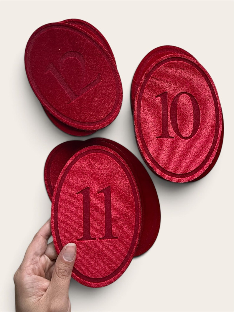 K&ouml;nnte beinhalten: Ein Set aus ovalen, roten Stoffanh&auml;ngern mit den gepr&auml;gten Zahlen 10, 11 und 12. Jeder Anh&auml;nger hat eine strukturierte Oberfl&auml;che mit einem erhabenen Rand und einer mittig platzierten Ziffer. Die Anh&auml;nger sind gestapelt und gruppiert angeordnet.