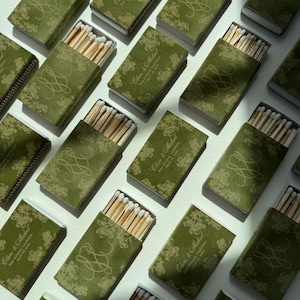 May include: A collection of rectangular matchboxes with a dark olive green velvet-like texture. Each box features a gold-toned floral pattern, a decorative monogram, and the printed text Claire & Michael, New Orleans, Louisiana, April 19, 2024.