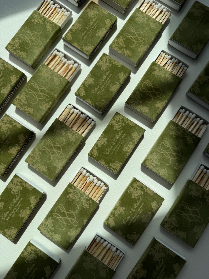 May include: A collection of rectangular matchboxes with a dark olive green velvet-like texture. Each box features a gold-toned floral pattern, a decorative monogram, and the printed text Claire & Michael, New Orleans, Louisiana, April 19, 2024.
