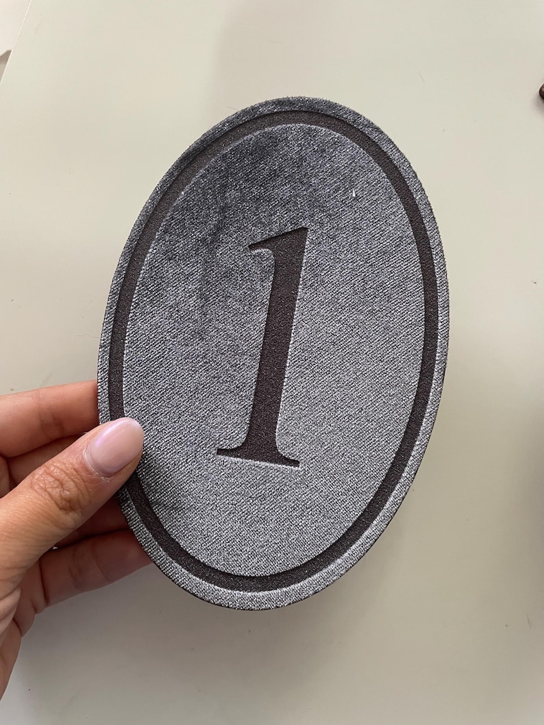 K&ouml;nnte beinhalten: Ein ovaler Aufn&auml;her mit einem dunkelgrauen, strukturierten Stoffhintergrund und einer markanten schwarzen Eins in der Mitte. Das Design umfasst einen d&uuml;nnen, dunklen kreisf&ouml;rmigen Rand, der die zentrale Ziffer einrahmt und f&uuml;r einen klaren Look sorgt.