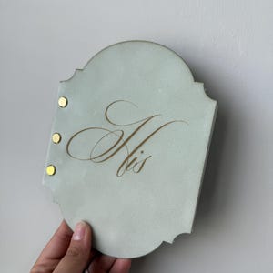 May include: A light blue, scalloped-edge plaque with the word "His" in gold script. Three gold rivets secure the plaque. The plaque is held by a hand against a neutral background.