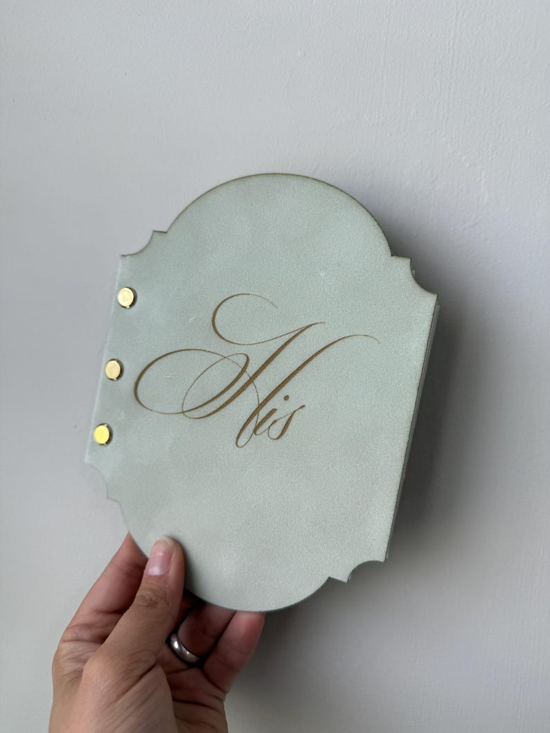 May include: A light blue, scalloped-edge plaque with the word "His" in gold script. Three gold rivets secure the plaque. The plaque is held by a hand against a neutral background.