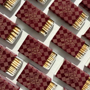 May include: Multiple rectangular matchboxes arranged in a grid pattern. Each box features a dark red and light brown checkered pattern with a central ornate monogram logo. The boxes are partially open, revealing rows of wooden matches with white tips.