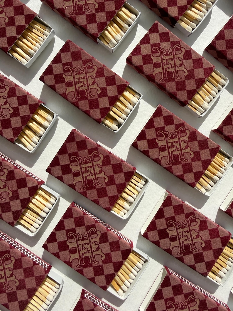 May include: Multiple rectangular matchboxes arranged in a grid pattern. Each box features a dark red and light brown checkered pattern with a central ornate monogram logo. The boxes are partially open, revealing rows of wooden matches with white tips.