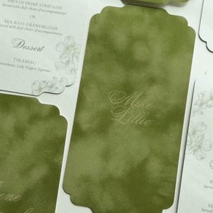 May include: A collection of rectangular paper cards with scalloped edges, featuring a mottled olive green finish and white cards with floral line art. The green cards display names like Mike and Lillie in script, while white cards list menu items.