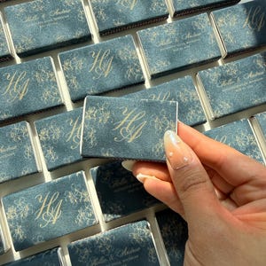 May include: A collection of rectangular matchboxes featuring a dark blue textured cover with a gold floral pattern and the monogram HA. Some boxes display the text Hollie & Andrew, Rochester Hills, Michigan, May 23, 2026 in gold lettering.