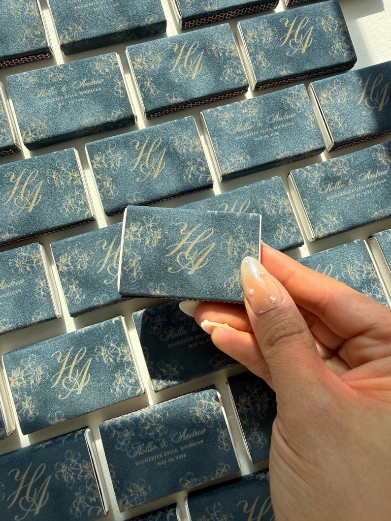 May include: A collection of rectangular matchboxes featuring a dark blue textured cover with a gold floral pattern and the monogram HA. Some boxes display the text Hollie & Andrew, Rochester Hills, Michigan, May 23, 2026 in gold lettering.