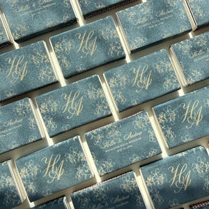 May include: A collection of rectangular matchboxes featuring a dark blue textured surface with gold foil printing. The designs alternate between a large monogram of the letters HA and the text Hollie & Andrew, Rochester Hills, Michigan, May 25, 2024.