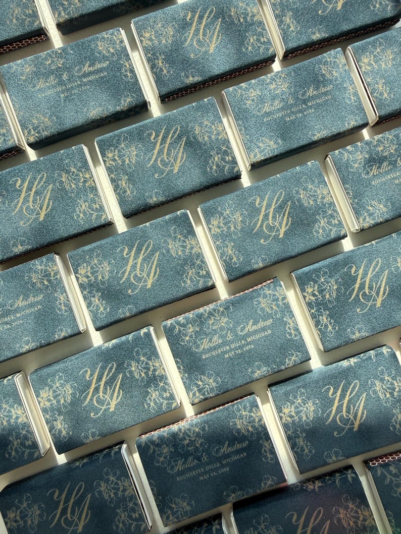 May include: A collection of rectangular matchboxes featuring a dark blue textured surface with gold foil printing. The designs alternate between a large monogram of the letters HA and the text Hollie & Andrew, Rochester Hills, Michigan, May 25, 2024.