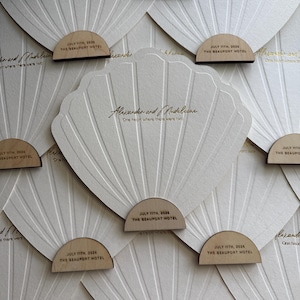 Op de afbeelding: Een verzameling witte, schelpvormige papieren waaiers met goudkleurige tekst Alexander and Madeline, One heart where there were two. Elke waaier heeft een halfrond houten handvat met de opdruk JULY 11TH, 2024 THE BEAUPORT HOTEL.