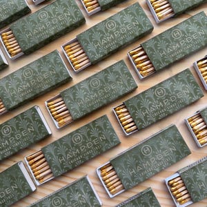 May include: Multiple rectangular matchboxes with a sage green cover featuring a repeating fan pattern and the text Hampden Chicago Since 1922. Each box is partially open, revealing a row of wooden matches with yellow-coloured tips inside.