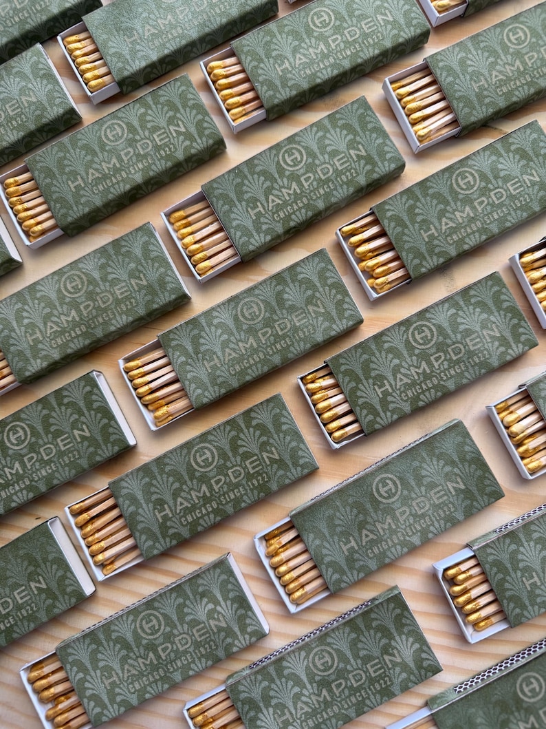May include: Multiple rectangular matchboxes with a sage green cover featuring a repeating fan pattern and the text Hampden Chicago Since 1922. Each box is partially open, revealing a row of wooden matches with yellow-coloured tips inside.