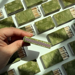 May include: A collection of rectangular matchboxes with olive green covers featuring a gold floral pattern, a gold monogram, and the text Claire & Michael, New Orleans, Louisiana, April 18, 2024. Each box contains wooden matches with white tips.