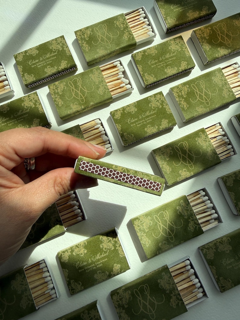 May include: A collection of rectangular matchboxes with olive green covers featuring a gold floral pattern, a gold monogram, and the text Claire & Michael, New Orleans, Louisiana, April 18, 2024. Each box contains wooden matches with white tips.