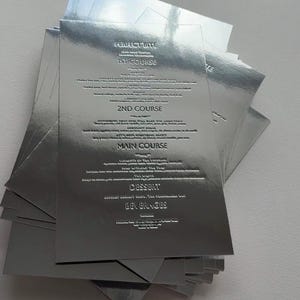 Op de afbeelding: Een stapel zilverkleurige menu's met reliëftekst. De menu's zijn in een waaier gerangschikt, met de tekst zichtbaar op het bovenste menu. Het menu bevat secties voor gangen zoals "Perfect Bite", "1st Course", "2nd Course", "Main Course", "Dessert" en "Dranken".