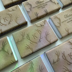May include: A collection of rectangular matchboxes featuring a tan, suede-like texture with embossed antler patterns. Some boxes display the letter K, while others show the text Kulasi, 1373 Gato Del Sol Avenue, Austin, Texas 78737 in dark brown ink.