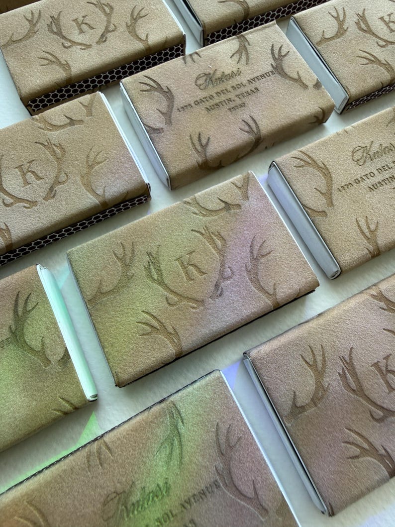 May include: A collection of rectangular matchboxes featuring a tan, suede-like texture with embossed antler patterns. Some boxes display the letter K, while others show the text Kulasi, 1373 Gato Del Sol Avenue, Austin, Texas 78737 in dark brown ink.