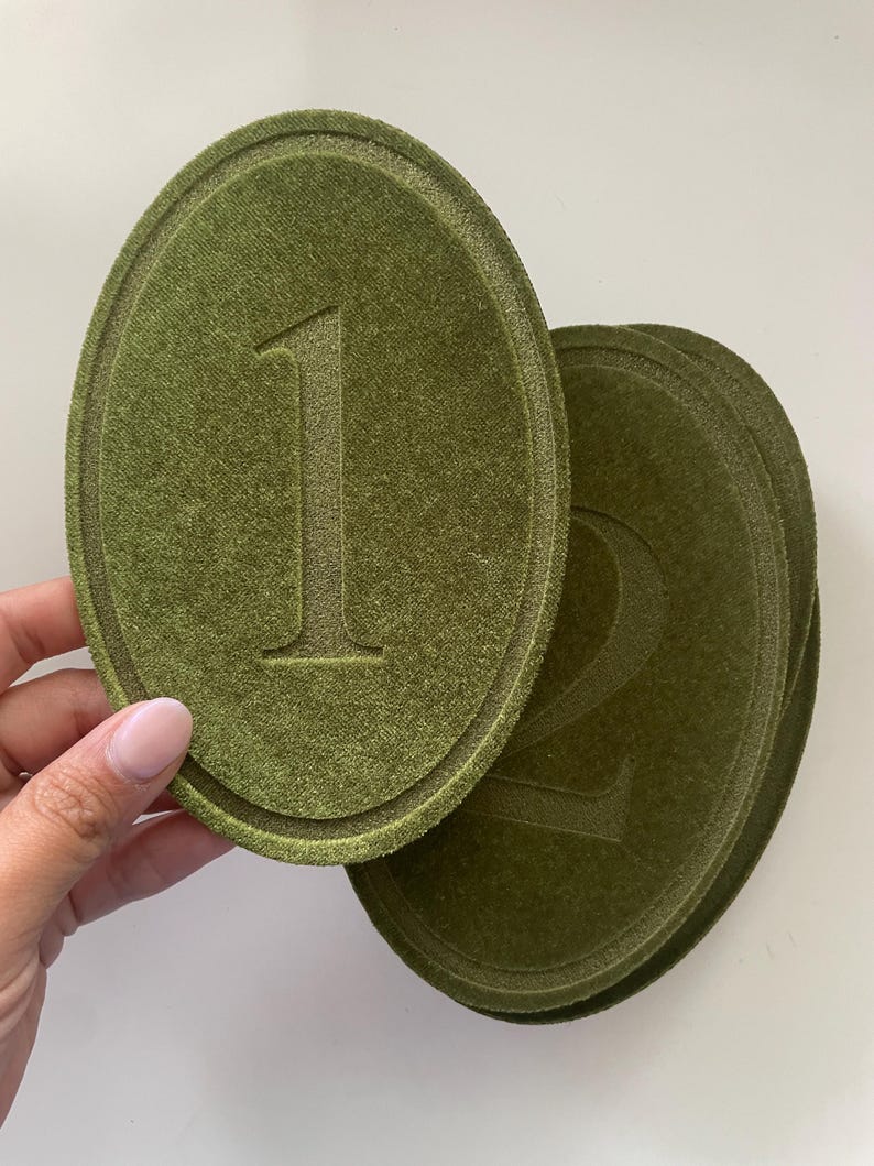 K&ouml;nnte beinhalten: Ein Set aus ovalen, olivgr&uuml;nen Untersetzern oder Dekoschildern aus Samt. Jedes St&uuml;ck ist in der Mitte mit einer eingepr&auml;gten Zahl versehen, wobei die Eins auf dem obersten St&uuml;ck und die Zwei auf dem darunter liegenden St&uuml;ck deutlich zu sehen ist.
