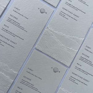 May include: A white menu card with a seashell logo and a list of food options for a wedding or special event. The menu includes Truffled Lobster Mac & Cheese, Seasonal Waldorf Salad, Line caught Grouper & Seasonal Succotash, and an assortment of locally made desserts and ice cream.